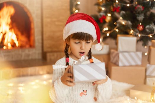 Merry Christmas! Happy child with gift box at home in decorated living room, looks with surprised facial expression on her present, keeps mouth widely opened, wearing santa hat, kid sits near tree.