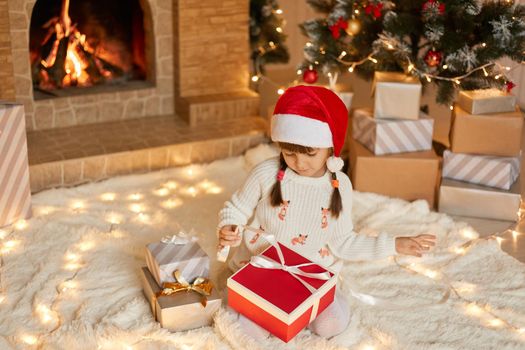 Cute little girl opening Christmas gift at home while sitting on soft carpet on floor, looking at box, pulls ribbon to open present, dresses white pullover and red santa hat.