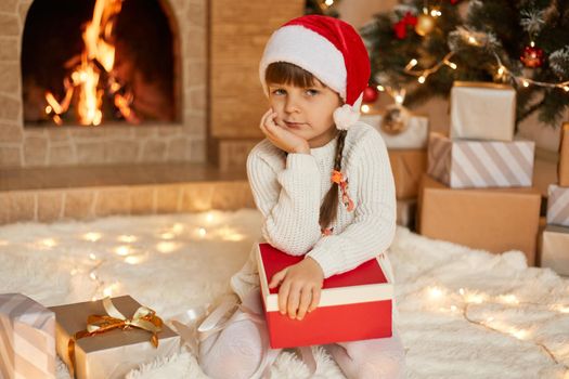 Child girl sitting near xmas tree and fireplace on Christmas Eve, holding gift box on knees, looking at camera with pensive expression, keeps hand under chin, wearing santa hat and jumper.