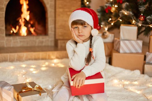 Pensive little girl thinking what to give for her family, kid wearing Santa Claus hat and white jumper, keeping palm under chin, having thoughtful facial expression, posing in festive room.