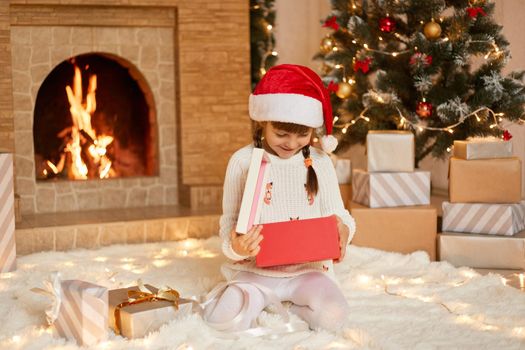 Happy small girl opening Christmas present box, looks inside with smile, being happy to get such gift, wearing white pullover and santa claus hat, sitting on floor in festive room with xmas decoration