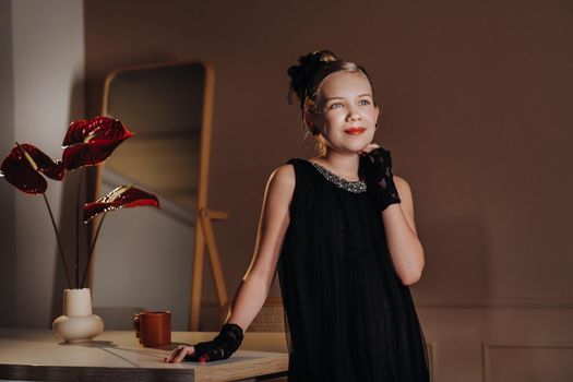 A stylish little girl in a black dress stands in the interior near the table.