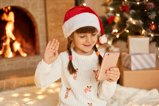 Happy child looking at smart phone screen and waving hand, video calling friends or relatives, stays at home for Christmas holidays, wearing white sweater and festive santa claus hat near fireplace.
