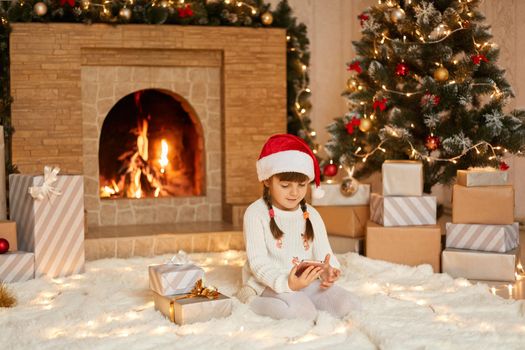 Cute girl with phone in hands sitting in living room on floor, dresses festive hat and casual attire, playing game on smartphone on christmas eve, posing near xmas tree and fireplace on soft carpet.