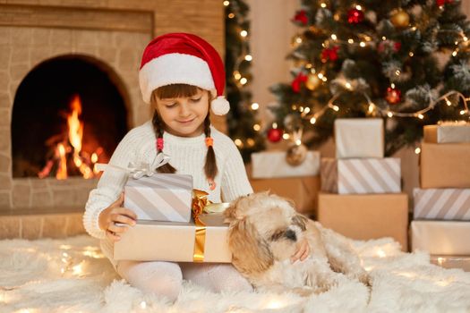 Happy small girl in santa hat have christmas, sitting on floor near xmas tree with gift boxes, having pleasant facial expression, wearing casual attire, posing in festve room.