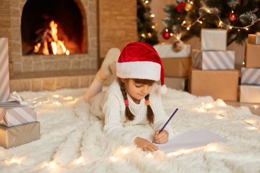 Little cute female child lying on floor on carpet and writing letter to Santa Claus, looking at empty paper and think, posing against present boxes, xmas tree and fire places.