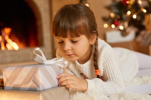 Pensive child with christmas present at home, female kid looks at gift box with ribbon and dreaming to open it, little girl with pigtails posing in living room near xmas tree and fireplace.