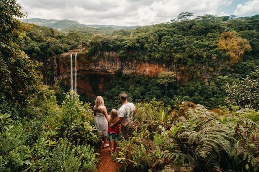 a family, a man, a woman and a daughter, stand on the edge of a cliff near a waterfall in Chamarel Park on the island of Mauritius.A couple with their daughter in the jungle of the island of Mauritius looks at a large waterfall falling down