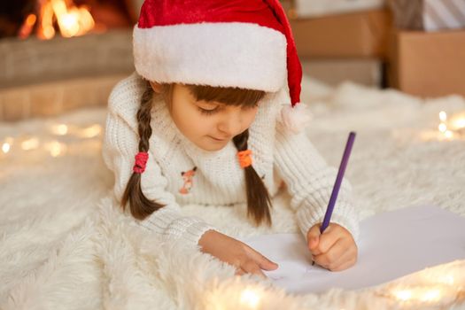 Little girl in red christmas hat writes letter to Santa Claus, posing on floor on soft carpet, child with two pigtails holding pencil in hands, kid with concentrated look dreams about xmas present.