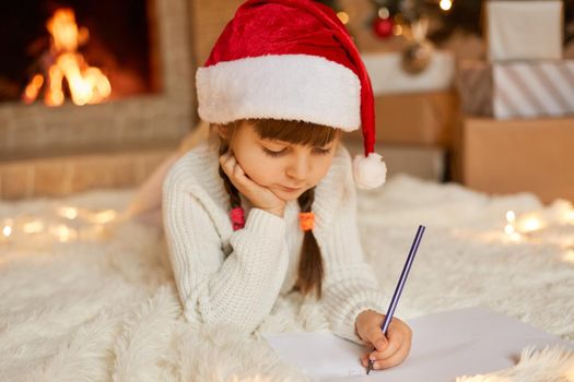 Little concentrated girl wearing white jumper and re santa hat posing on floor while drawing, keeping hand under chin, looking on white paper, being photographed against fireplace.