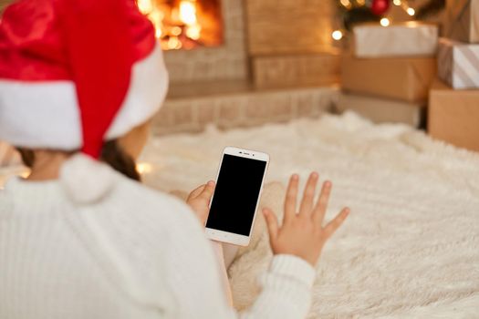 Little girl looks at blank phone screen and waving hand, back view of female kid wearing santa hat and white jumper, sitting on soft carpet on floor near fireplace .