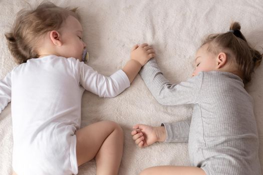 Childhood, sleep, relaxation, family, lifestyle concept - two young children 2 and 3 years old dressed in white and beige bodysuit sleep on a beige and white bed at lunch holding hands top view
