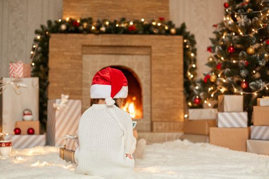 Child looking at smart phone or tablet screen while sitting near fireplace on floor, posing backwards, wearing white sweater and santa hat, posing in living room with new year decoration.