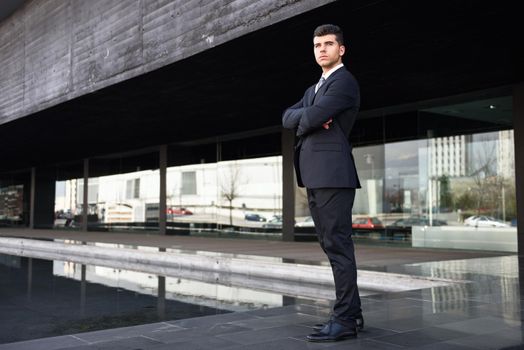 Young businessman near a modern office building wearing black suit and tie. Man with blue eyes