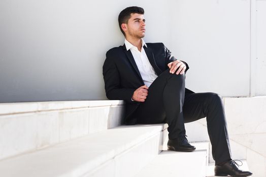 Young businessman near a modern office building wearing black suit and white shirt sitting on the floor. Man with blue eyes in urban background