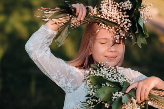 A beautiful nine-year-old blonde girl with long hair in a long white dress, holding a bouquet of lilies of the valley flowers, walking in nature in the Park.Summer, sunset