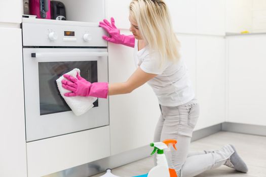 Woman in the kitchen is smiling and wiping dust using a spray and a duster while cleaning her house, close-up