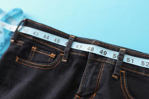 Close-up of upper part of denim trousers, jeans with blue measure tape instead of belt. Individual clothes sew in atelier. Healthy lifestyle, dieting, weight loss, tailoring concept