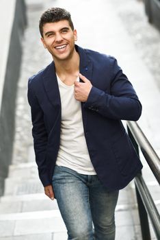 Handsome young man smiling with blue eyes in the street. Model of fashion walking in urban background wearing white t-shirt, jeans and blue jacket