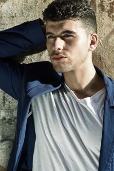 Close-up portrait of a young man with blue eyes posing near a wall. Model of fashion in urban background wearing white t-shirt and blue jacket