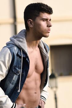 Good looking young man with muscular body in the street. Model of fashion in urban background wearing jeans and blue jacket