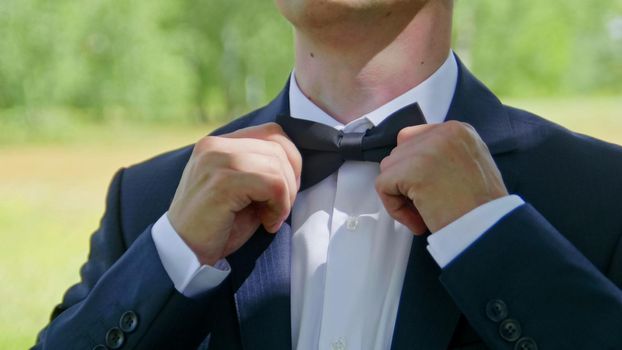 The groom puts on a bow tie from the front, outdoor, closeup.