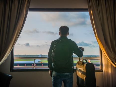 A young man watches the airplane take off from the window of his hotel room
