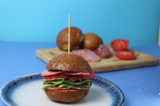 sandwich, burger with buckwheat brown bun vegetables pieces of tomatoes cucumbers on a blue plate on a blue background are the ingredients for making a burger vegetables sausage ham.