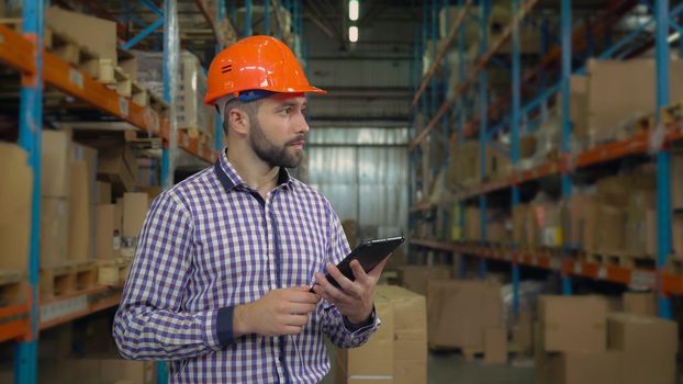 Manager working at warehouse. Handsome young man wearing casual shirt and hard hat using digital tablet entering data. Worker counting box for delivery.