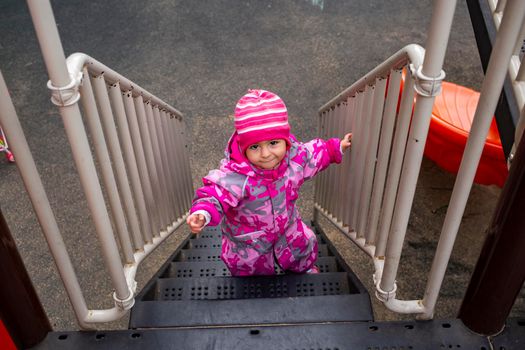 adorable toddler climbs the stairs on the playground. toddler baby dressed in snowsuit. autumn or winter, cold season