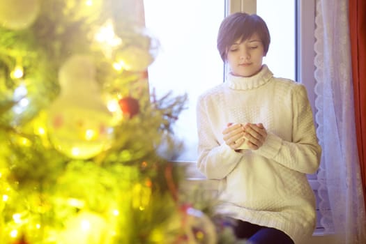 Young beautiful woman sitting home by the window with cup of hot coffee wearing knitted warm sweater. Christmas tree with decorations and lights in the room.