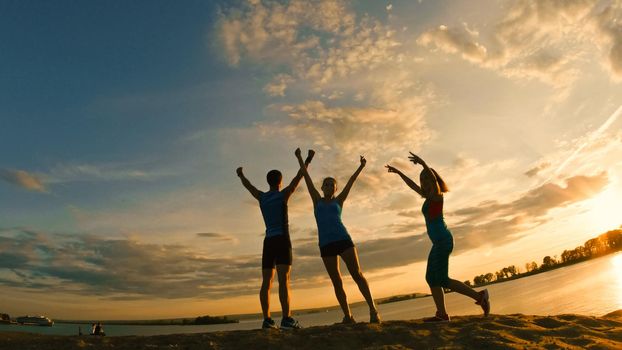 Group of young people - athletes - two girls and a guy are have triumph on the mountain, near river at dusk, silhouette