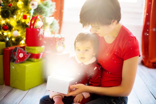 Young mother and her little daughter dressed as santa claus opening a magical Christmas gift by a Christmas tree in cozy living room in winter.