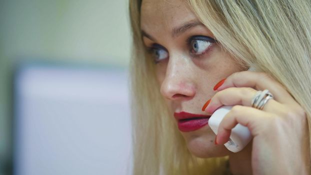 Young Blonde Woman in office talking on phone in front of the computer, extremely close up, telephoto
