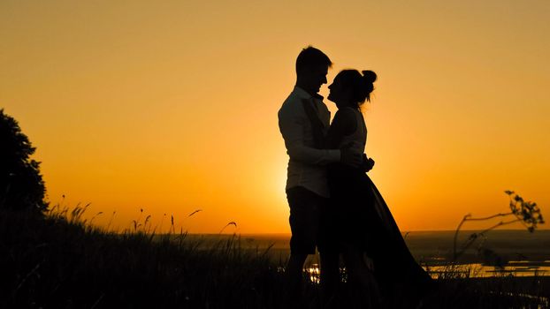 loving couple - brave young man and beautiful girl stands on high hill at sunset and have hugs and kiss, silhouette, telephoto shot