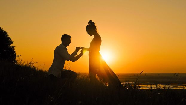 Romantic Silhouette of Man Getting Down on his Knee and Proposing to Woman on high hill - Couple Gets Engaged at Sunset - Man Putting Ring on Girl's Finger, wide angle, slow motion