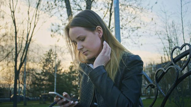 Cute young girl uses phone and straightens hair sitting on the bench at sunset, horizontal