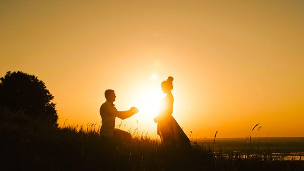 Romantic Silhouette of Man Getting Down on his Knee and Proposing to Woman high hill - Couple Gets Engaged at Sunset - Putting Ring Girl's Finger, telephoto