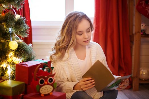 Young beautiful woman sitting on floor near christmas tree and reading christmas postcard.