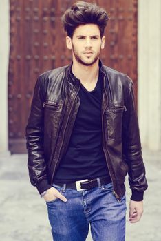 Portrait of a young handsome man, model of fashion, with modern hairstyle in urban background wearing blue jeans and leather jacket