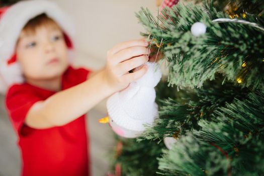 Cute little boy of about five year with blue eyes in a Santa Claus hat decorating a Christmas tree. .