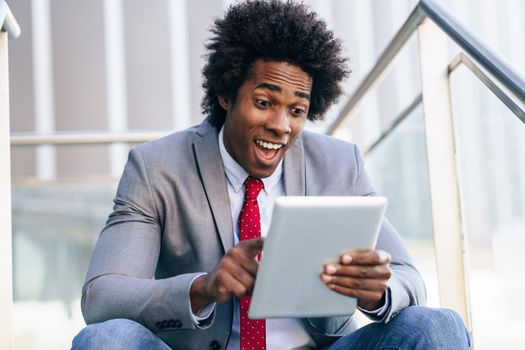 Black Businessman using a digital tablet sitting near an office building. Man with afro hair and surprise expression