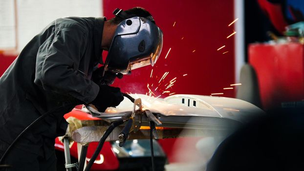 Welding industrial: worker in helmet repair detail in car auto service, telephoto