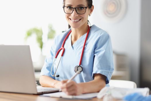 Portrait of woman therapist posing in clinic office. Smiling woman physician sitting at table in consulting room. Cheerful practitioner working with documents. Medicine and healthcare concept