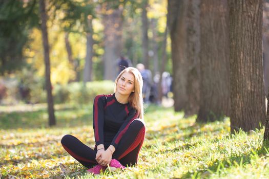 Young fitness beauty girl sitting cross-legged at workout outdoor near trees, telephoto
