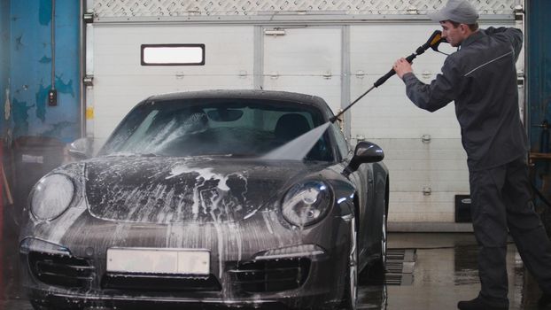 Mechanic in car service is washing sportcar in the suds by water hoses, telephoto