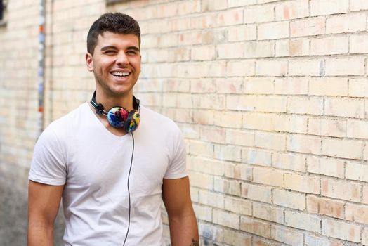 Portrait of young man in urban background smiling with headphones. Wearing white t-shirt near a brick wall