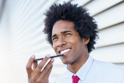 Black Businessman sending voice note with his smartphone near an office building. Man with afro hair.