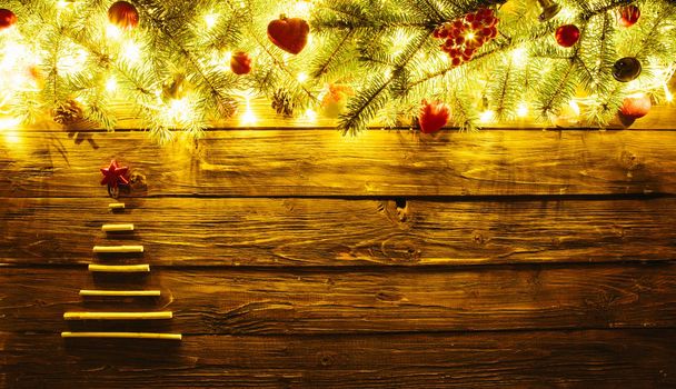 Blurred Christmas background, with fir branches, xmas tree from dry sticks, fairy lights and christmas decorations on brown wooden plank .
