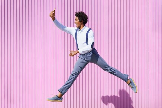 Black man, with afro hair, taking a selfie with his smartphone while jumping on a background of pink shutters.